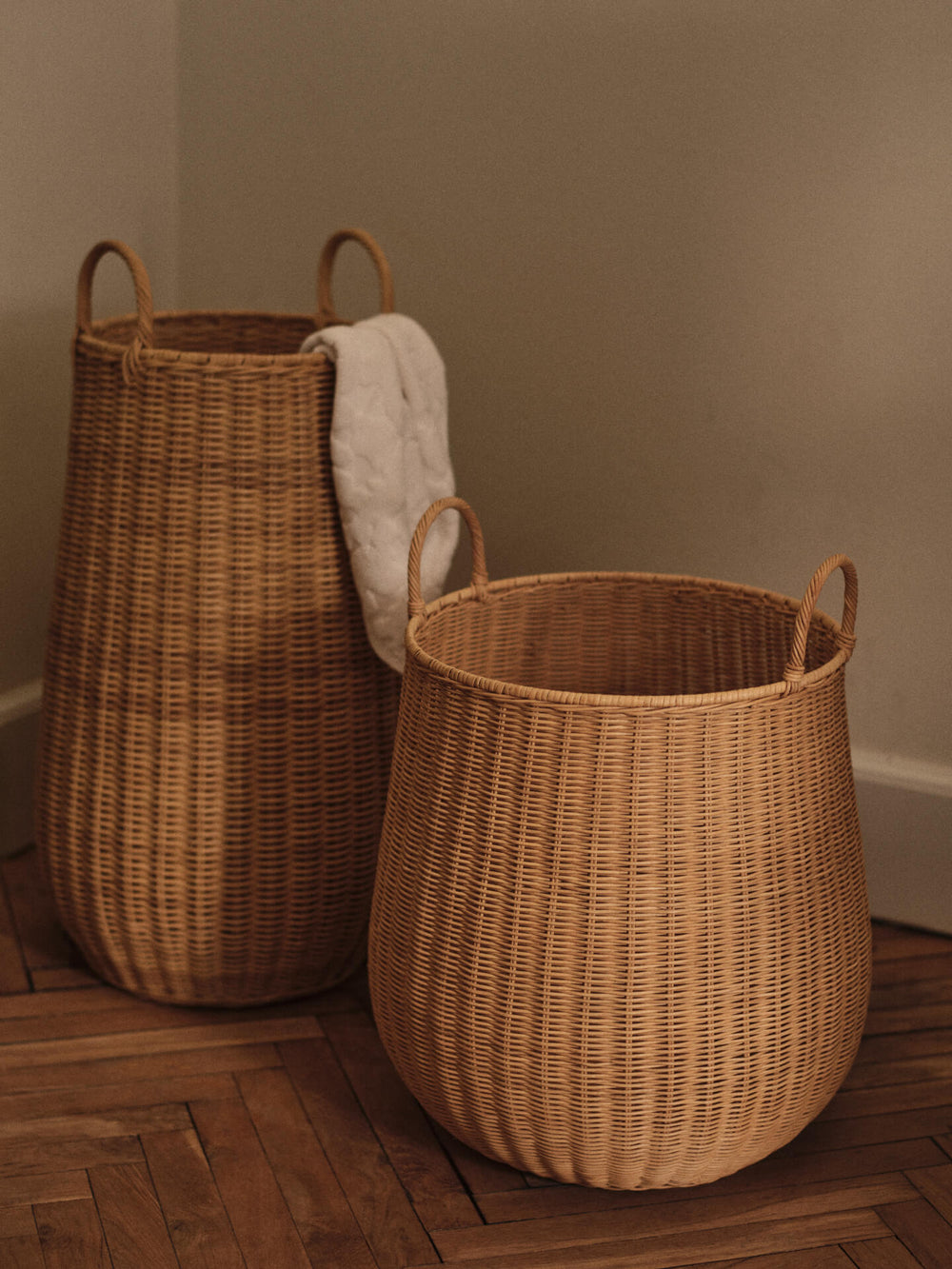 Two woven baskets with handles on a herringbone floor, a blanket draped over the taller one