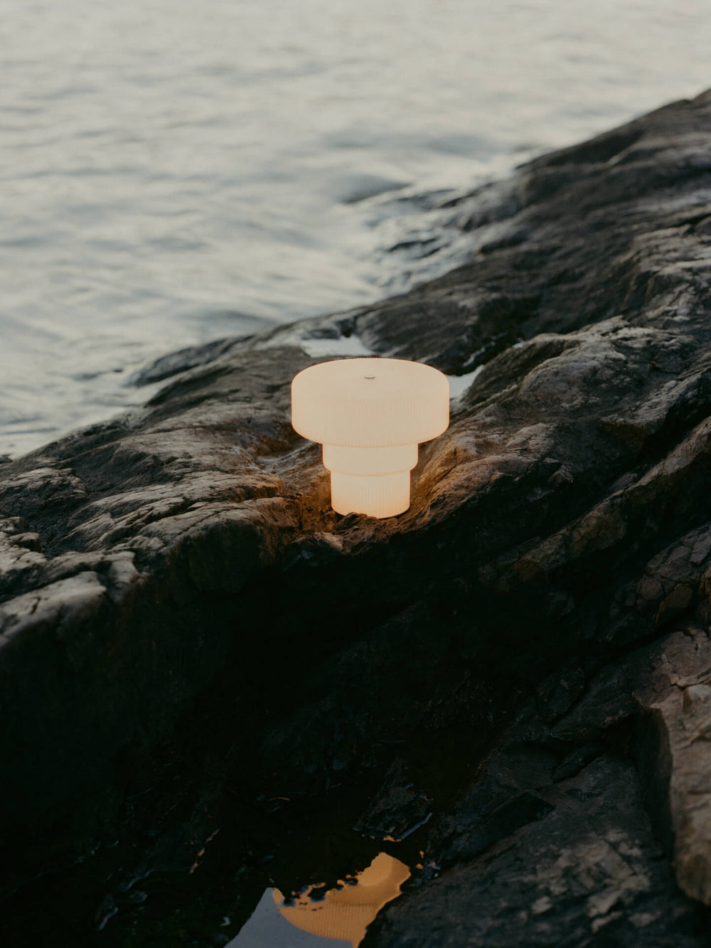 Lit portable lamp standing on dark coastal rocks at the water's edge at dusk