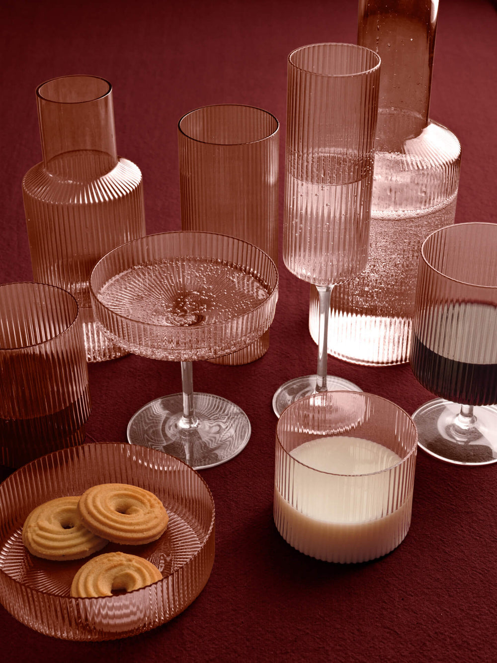 Ribbed blush glassware grouped on a dark red table, carafes, glasses, coupe and a dish of biscuits