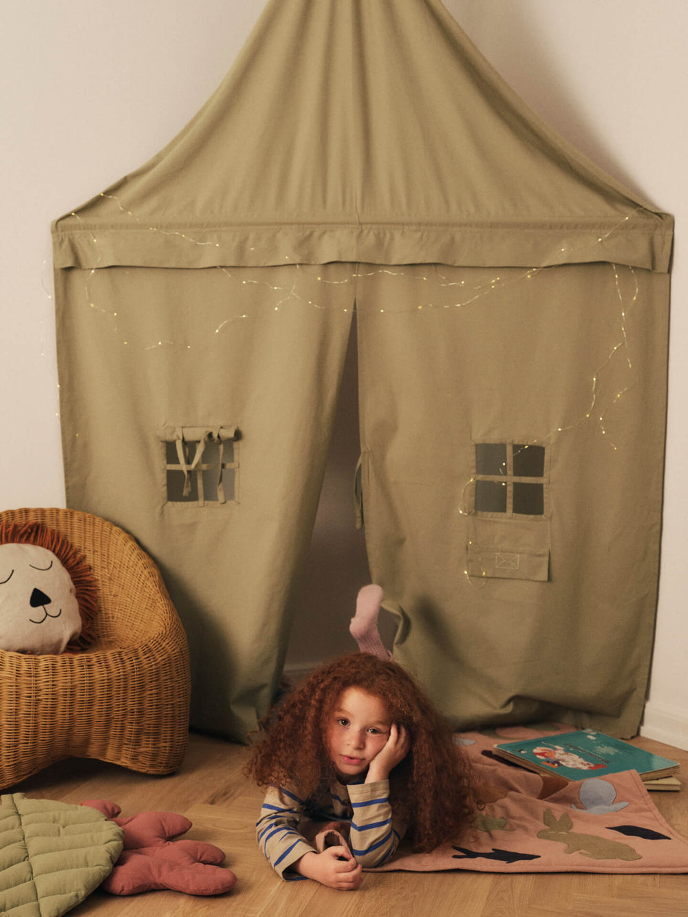 Child lying on a play mat in front of the corner curtain, with a lion cushion in a woven chair