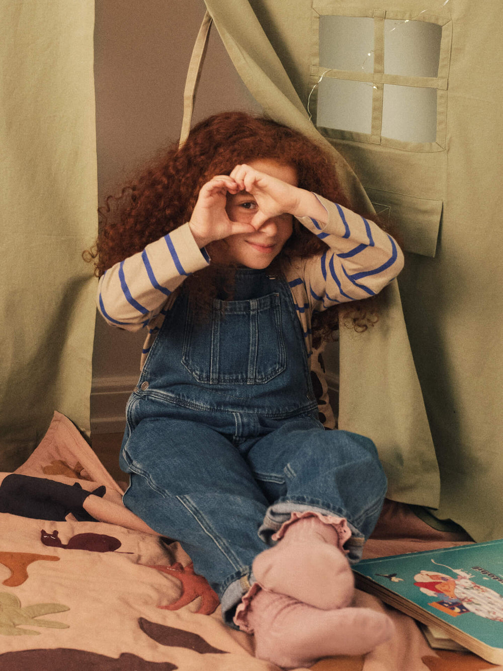 Child sitting under the corner curtain making a heart with their hands, patterned blanket and book alongside