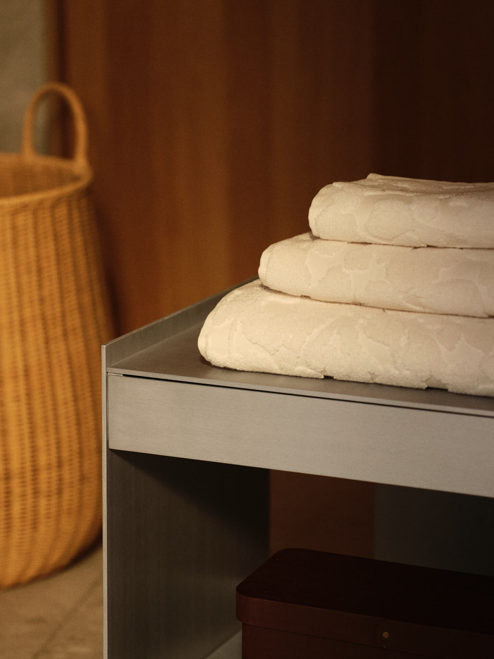 Stack of three off-white towels with tonal relief pattern on a bathroom bench, woven basket nearby