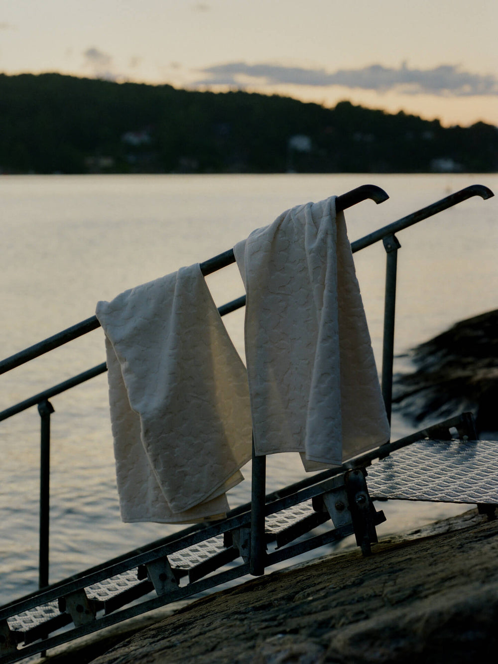 Two off-white bath towels hung over a railing at a waterside bathing ladder at dusk