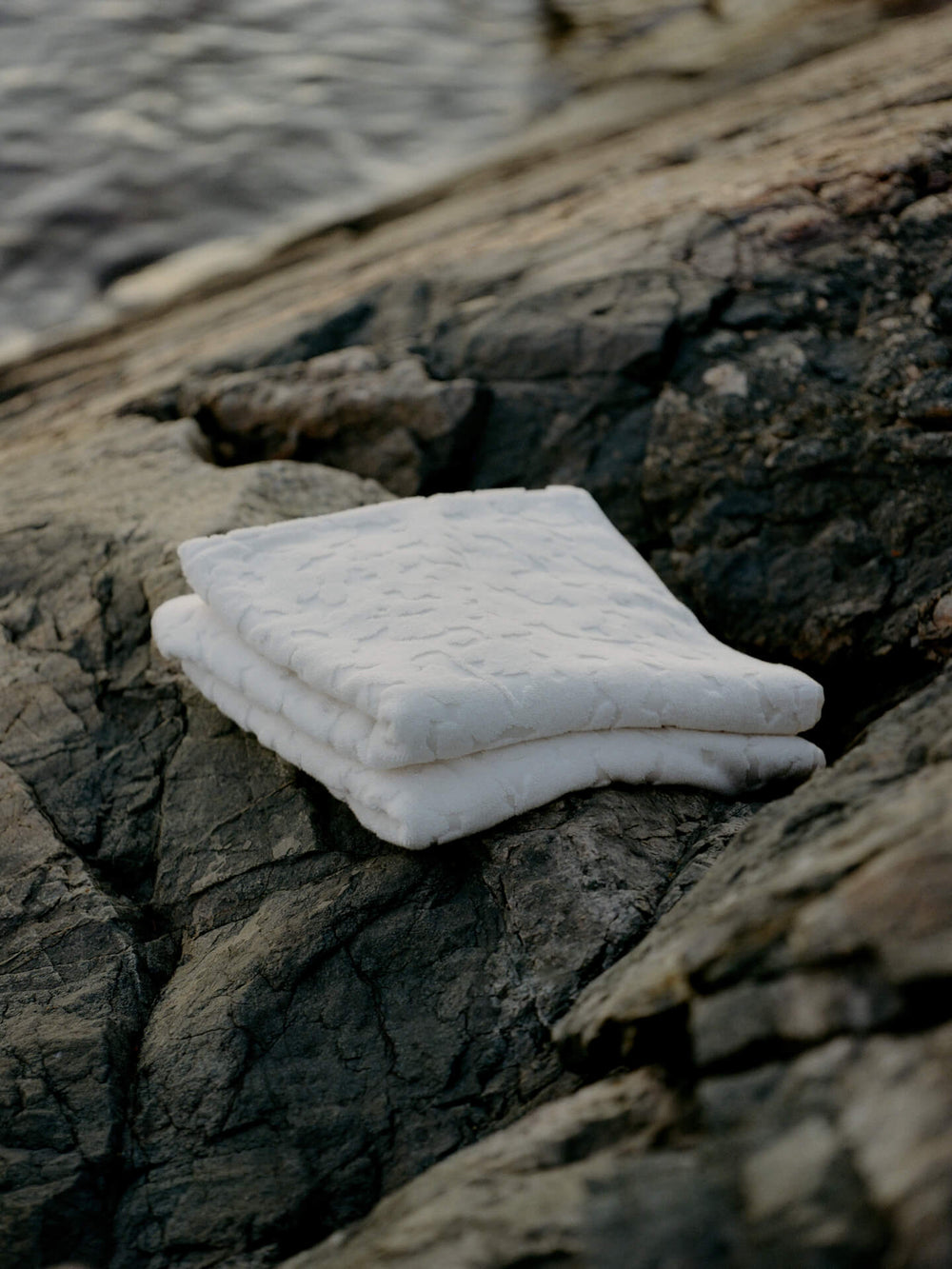 Two folded off-white towels with raised floral relief, stacked on coastal rock by the water