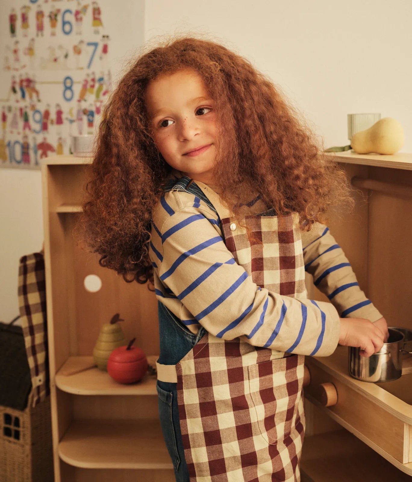 Kids Autumn Winter 2026 collection, child in a checked apron holding a small pot at a play kitchen
