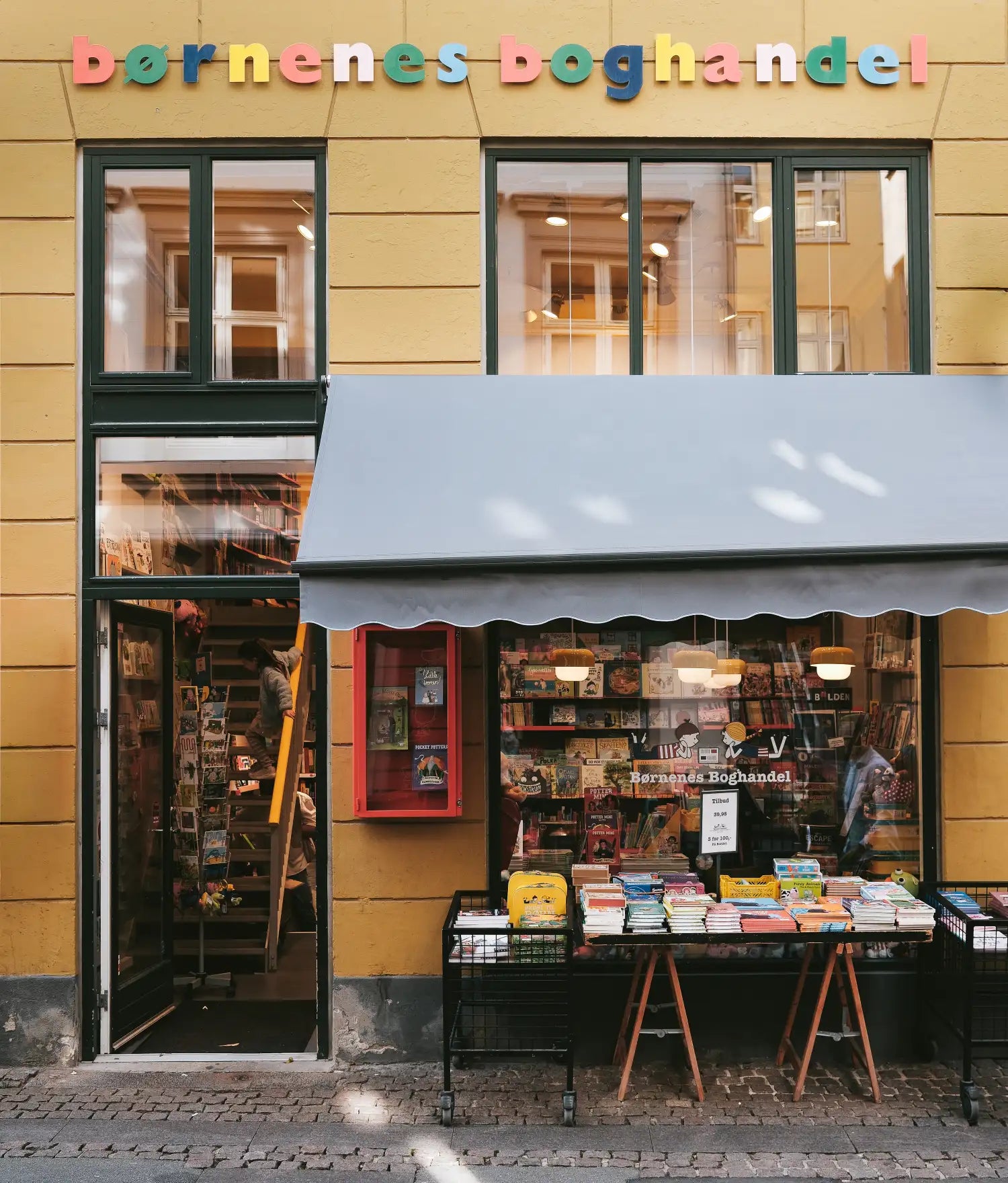 Yellow shopfront of the children's bookshop Børnenes Boghandel, awning and book tables on the pavement