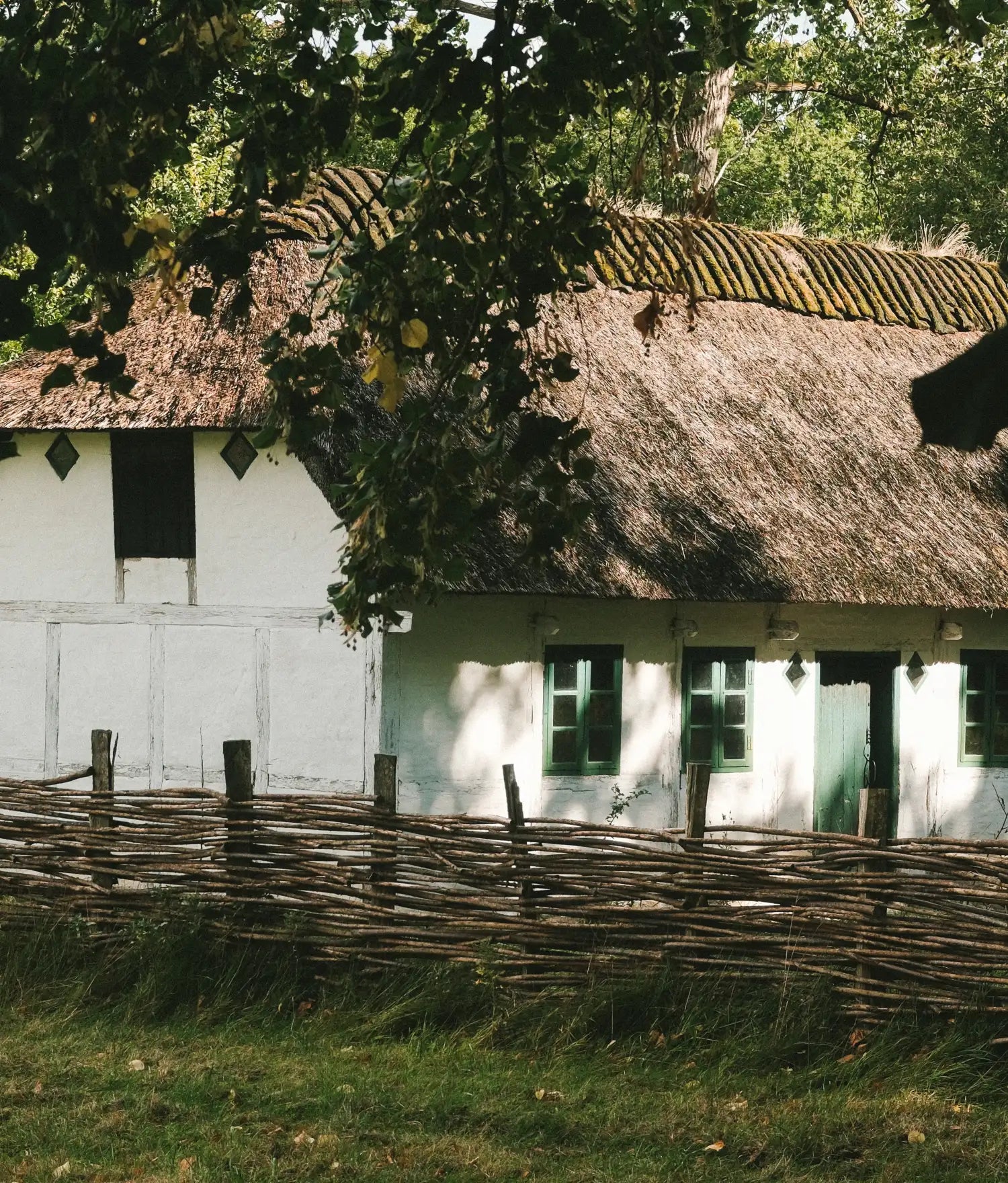 Whitewashed thatched cottage with green windows and door behind a woven branch fence