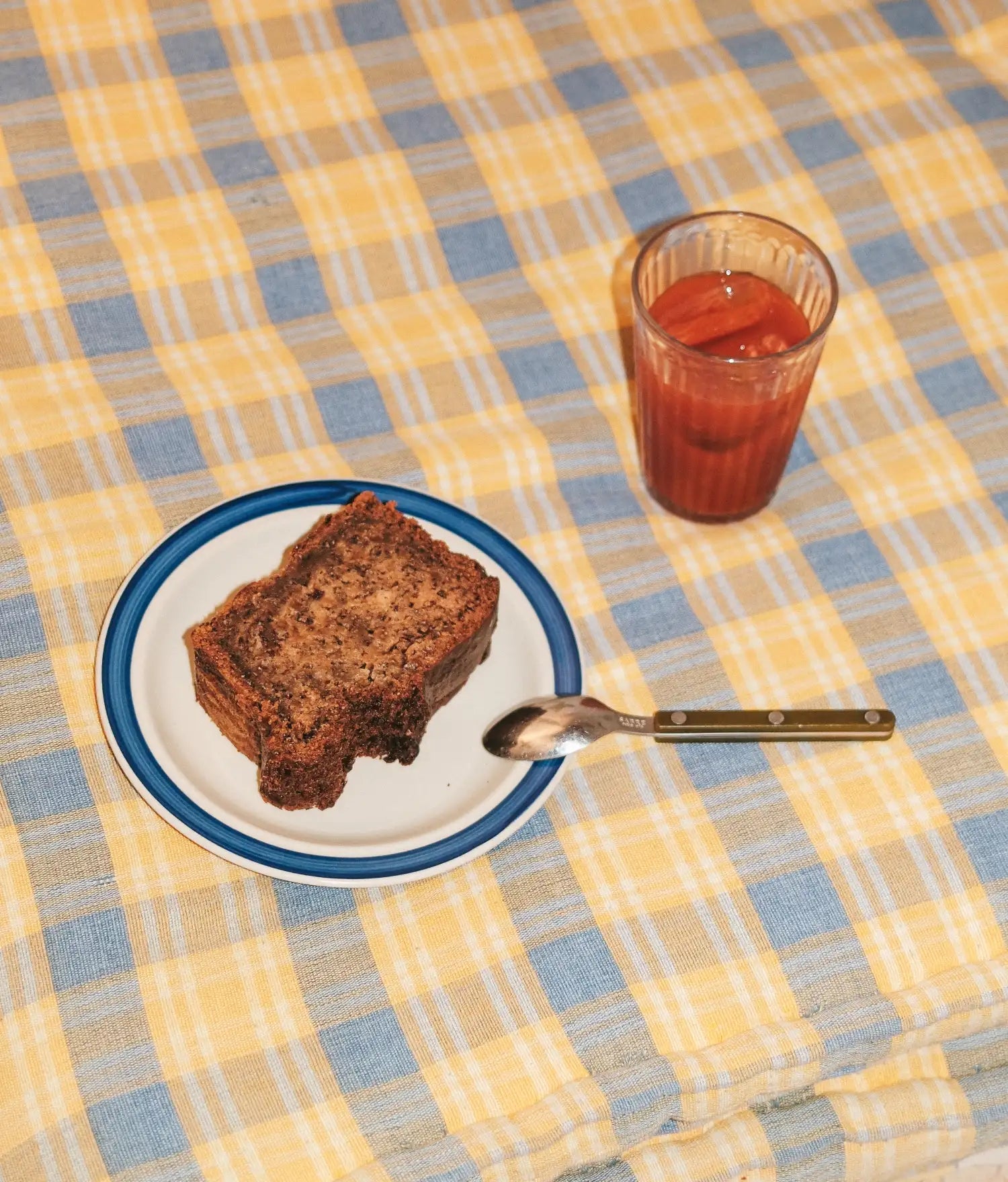 Checked tablecloth in yellow and blue with a slice of cake on a blue rimmed plate, spoon and iced drink