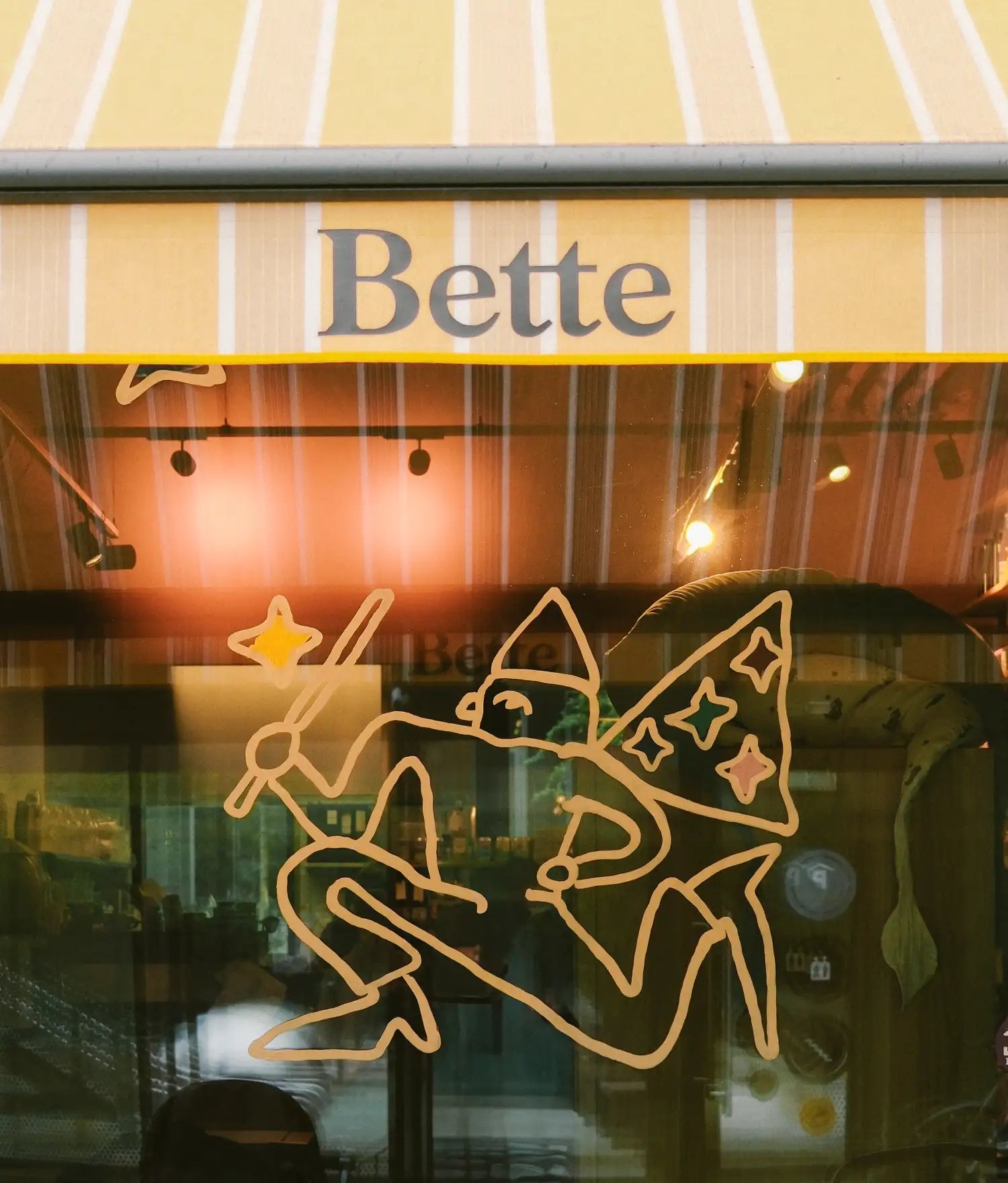 Shop window with striped yellow awning reading Bette, fairy line drawing on the glass